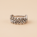 Anillo ajustable de acero con diseño de corona de laureles y espigas visto de frente sobre fondo beige